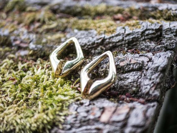 The DAYAK Triangle Keyhole Ear Hangers in Gold by Kaya Movement, designed for 00 gauge piercings, rest elegantly on a textured surface of moss-covered bark. The shiny gold brass reflects light beautifully, emphasizing their smooth surfaces and modern geometric design—perfect for those with stretched ear lobes.